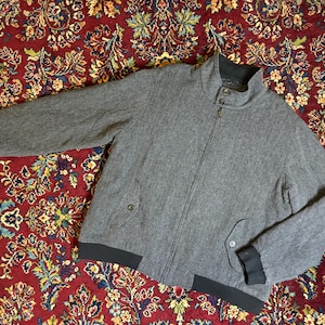 Puede incluir: Chaqueta gris con cuello, puños y dobladillo negros. La chaqueta tiene cierre de cremallera, dos bolsillos delanteros y cuello alzado. La chaqueta es de tejido texturizado y está sobre una alfombra estampada.