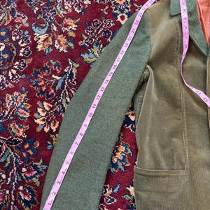 K&ouml;nnte beinhalten: Gr&uuml;ne Cordjacke mit gr&uuml;nem &Auml;rmel, gemessen mit einem rosa Ma&szlig;band. Der &Auml;rmel ist etwa 69 cm lang. Die Jacke hat ein braunes Futter und eine Tasche.
