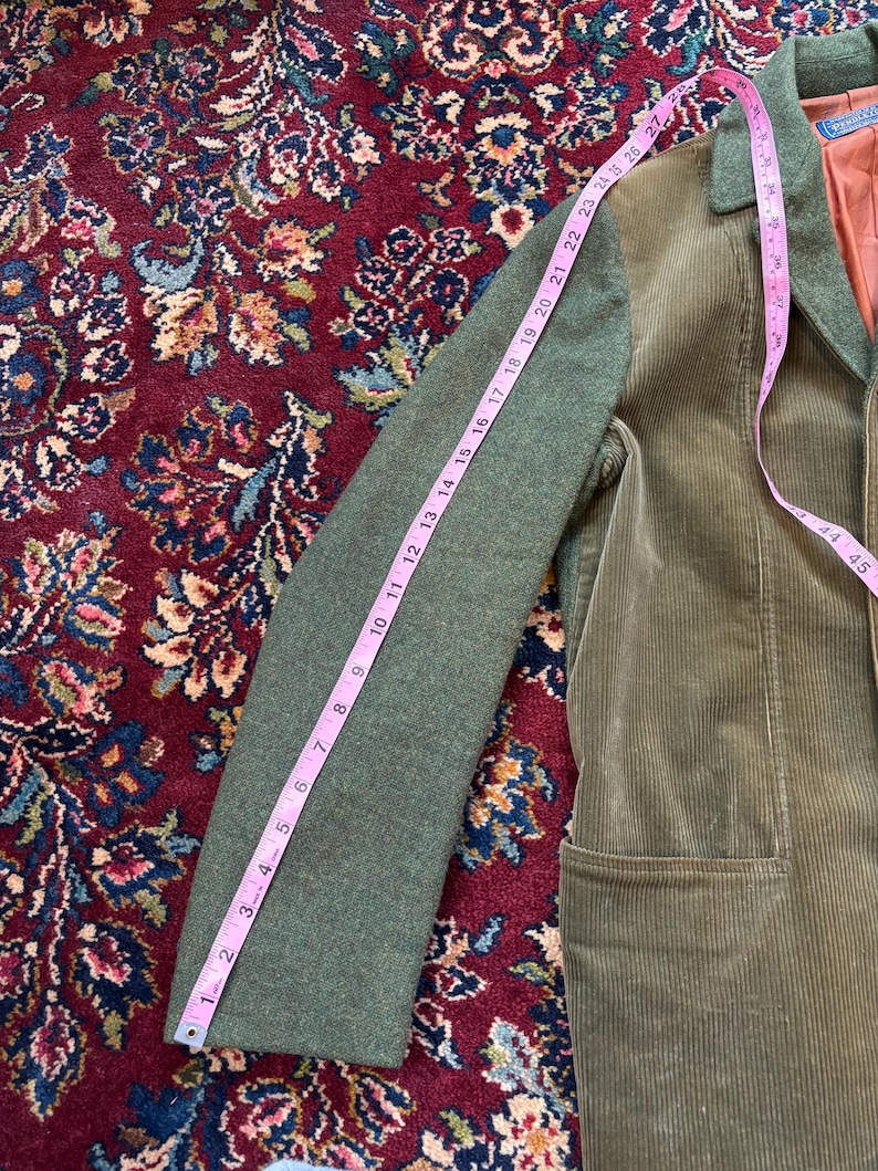 K&ouml;nnte beinhalten: Gr&uuml;ne Cordjacke mit gr&uuml;nem &Auml;rmel, gemessen mit einem rosa Ma&szlig;band. Der &Auml;rmel ist etwa 69 cm lang. Die Jacke hat ein braunes Futter und eine Tasche.