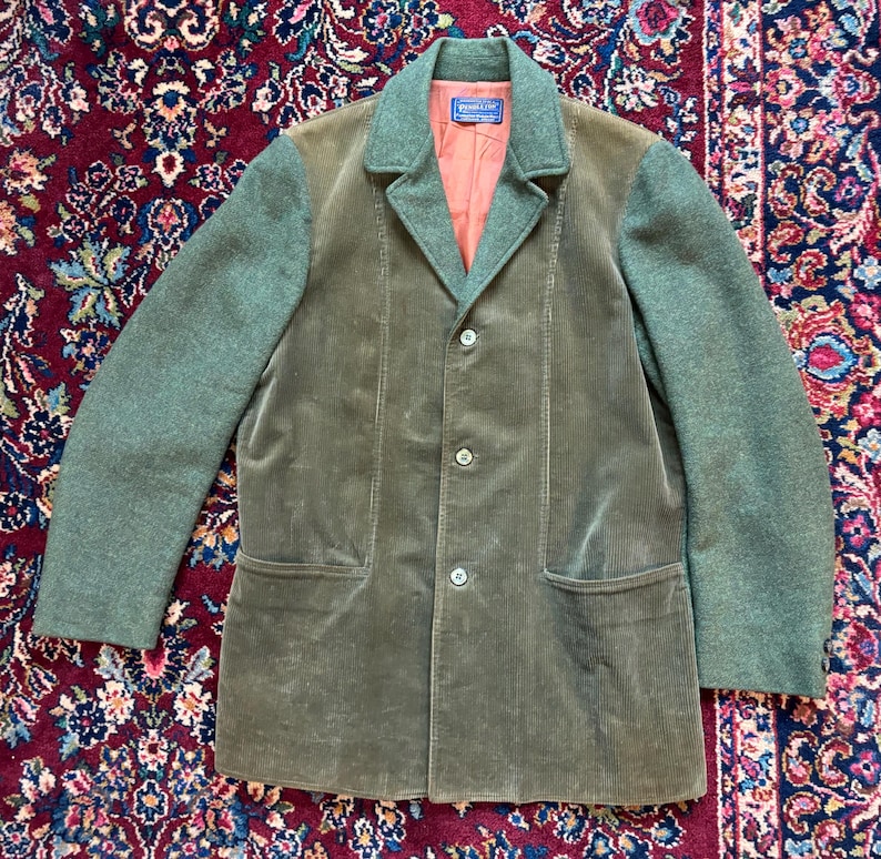 K&ouml;nnte beinhalten: Eine olivgr&uuml;ne Cordjacke mit kontrastierenden gr&uuml;nen Woll&auml;rmeln und Kragen. Die Jacke hat einen Dreiknopfverschluss, zwei Vordertaschen und ein pfirsichfarbenes Futter. Das Pendleton-Etikett ist am Kragen sichtbar.
