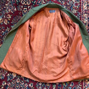 K&ouml;nnte beinhalten: Eine offene gr&uuml;ne Wolljacke mit pfirsichfarbenem Futter. Die Jacke liegt flach, wodurch das Innenfutter und das Etikett sichtbar werden. Die Au&szlig;enseite der Jacke ist einfarbig gr&uuml;n, das Futter ist glatt und einfarbig pfirsichfarben.