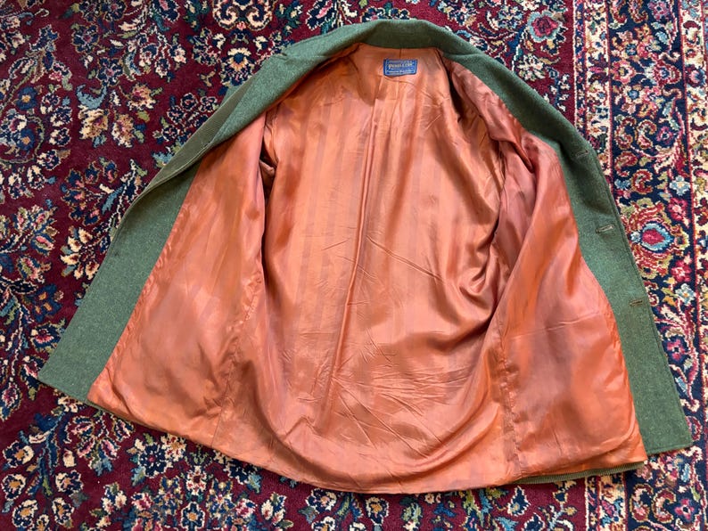 K&ouml;nnte beinhalten: Eine offene gr&uuml;ne Wolljacke mit pfirsichfarbenem Futter. Die Jacke liegt flach, wodurch das Innenfutter und das Etikett sichtbar werden. Die Au&szlig;enseite der Jacke ist einfarbig gr&uuml;n, das Futter ist glatt und einfarbig pfirsichfarben.