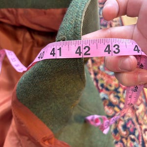 K&ouml;nnte beinhalten: Nahaufnahme eines gr&uuml;nen Wollmantels, der mit einem rosa Ma&szlig;band gemessen wird. Die Innenseite des Mantels ist rostorange. Das Band zeigt 104 cm an.