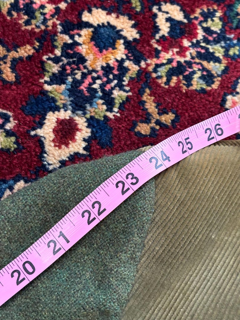 K&ouml;nnte beinhalten: Ein rosa Ma&szlig;band zeigt ein Ma&szlig; von 50,8 bis 66 cm &uuml;ber einem gr&uuml;nen Tweed-Stoff und einem Cordstoff. Die Stoffe sind olivgr&uuml;n und liegen auf einem rot, blau und beige gemusterten Teppich.