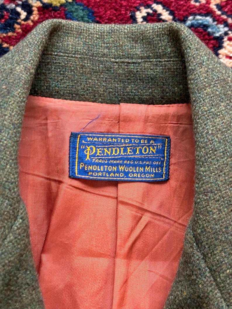 K&ouml;nnte beinhalten: Nahaufnahme eines gr&uuml;nen Tweed-Blazers mit pfirsichfarbenem Futter. Ein blaues Etikett im Inneren zeigt "Pendleton Woolen Mills, Portland, Oregon."