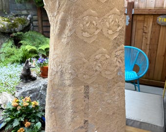 May include: A beige lace slip dress with spaghetti straps. The dress features a floral lace pattern and is displayed on a mannequin. The background includes outdoor elements like plants, a wooden fence, and a blue chair.