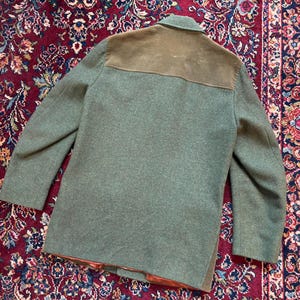 K&ouml;nnte beinhalten: Ein gr&uuml;ner Wollmantel mit einem braunen Wildleder-Schulterbesatz. Der Mantel hat ein klassisches Design mit Kragen und langen &Auml;rmeln. Der Mantel wird auf einem gemusterten Teppich pr&auml;sentiert.