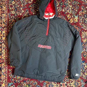 May include: Black Starter anorak jacket with a red interior lining and hood. The jacket features a red zipper, red stitching details, and the Starter logo in red across the chest. The jacket has a drawstring at the bottom and a Starter logo on the sleeve.