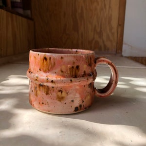 May include: A pink and brown ceramic mug with a speckled glaze. The mug has a wide, rounded base and a slightly flared lip. The handle is attached to the side of the mug and curves up to meet the lip.