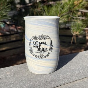 May include: A ceramic vase with a marbled blue and cream design. The vase features the text "let your light shine" in a floral wreath. The text is from Matthew 5:16.