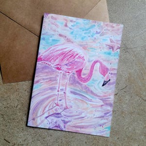 May include: A watercolor painting of a pink flamingo standing in water, with a brown envelope in the background. The flamingo has a curved neck and is surrounded by shades of pink, purple, and blue. The artwork is on a white card.
