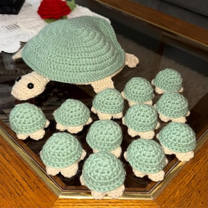 May include: A collection of handmade crochet turtles. The large turtle has a light green shell and a cream-colored head and legs. Several smaller turtles with green shells and cream-colored legs are arranged around the larger one.