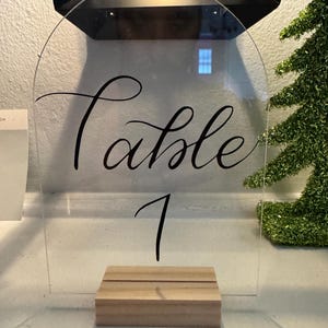 May include: Clear acrylic table number sign with a black calligraphy font that says "Table 1". The sign is on a wooden stand.