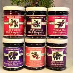 May include: Six glass jars of jam with white lids. The jars are labeled with the text "Genuine Oregon" and the names of the jams: "Old Fashion Red Raspberry", "Old Fashion Black Raspberry", "Wild & Rare Huckleberry", "Old Fashion Mountain Blackberry", "Old Fashion Coastal Cranberry", and "Old Fashion Marionberry".