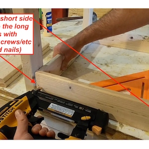 Op de afbeelding: Een persoon gebruikt een spijkerpistool om een ​​houten plank aan een andere houten plank te bevestigen. De planken maken deel uit van een grotere houten constructie. De tekst "Secure the short side aprons into the long side aprons with glue/nails/screws/etc (I used brad nails)" is zichtbaar in de afbeelding.