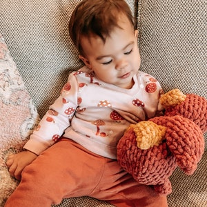 Op de afbeelding: Een jong kind zit op een bank en houdt een rood en geel gehaakt knuffeldier vast. Het kind draagt een roze longsleeve met een paddenstoelprint en een oranje broek.