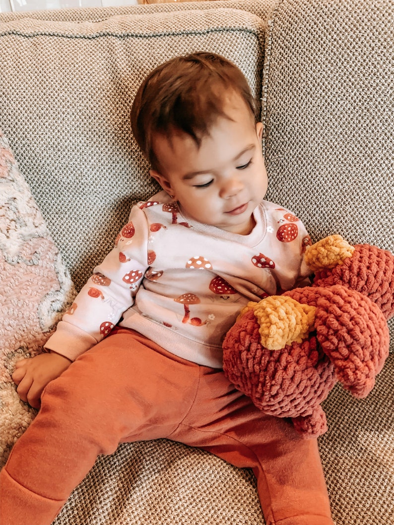 Op de afbeelding: Een jong kind zit op een bank en houdt een rood en geel gehaakt knuffeldier vast. Het kind draagt een roze longsleeve met een paddenstoelprint en een oranje broek.