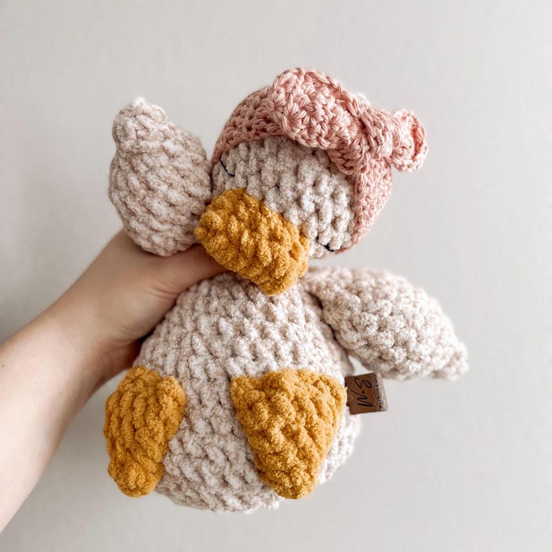 Op de afbeelding: Een gehaakt pluche speelgoed eendje met een roze gehaakte strik op zijn kop. De eend heeft een beige lichaam met gele gehaakte accenten op zijn snavel en voeten. Het speelgoed heeft een bruin label met de tekst "M&S" erop.