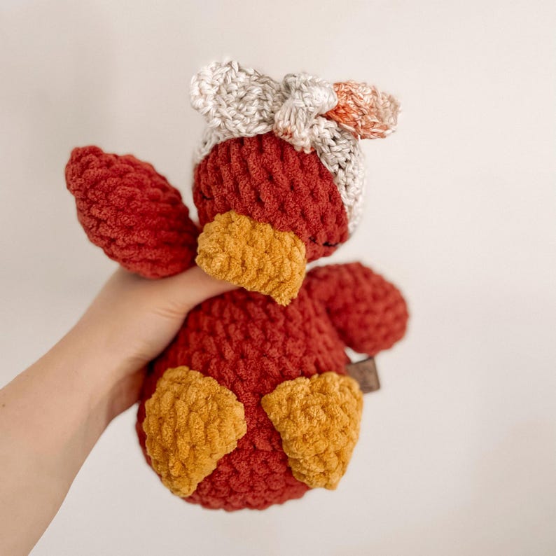 Op de afbeelding: Een gehaakt pluche speelgoed in de vorm van een vogel. De vogel is rood met gele accenten en heeft een witte en oranje gehaakte hoed.
