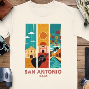 Könnte beinhalten: Cremefarbenes T-Shirt mit einem farbenfrohen San Antonio, Texas-Design. Die Grafik zeigt Illustrationen des Alamo, einer Gitarre und einer Flussszene mit dem Text "SAN ANTONIO TEXAS" in brauner Schrift.
