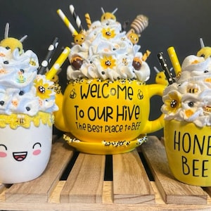 May include: Three yellow ceramic mugs with white whipped cream and bee decorations. The mugs are decorated with the text "Welcome to our Hive The Best Place to Bee", "Honey Bee", and a smiley face.
