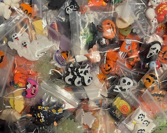 May include: Assortment of Halloween-themed charms and embellishments in various colours and designs, including ghosts, pumpkins, bats, and skulls. The charms are packaged in small plastic bags.