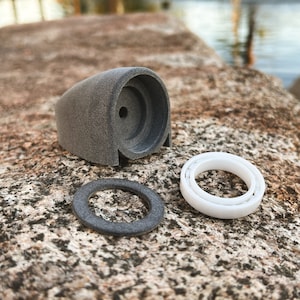 May include: A gray 3D printed part with a round opening, a white ring, and a black ring. The parts are on a gray textured surface.