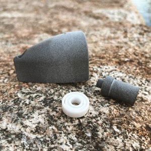 May include: A disassembled gray component with a white bearing. The gray parts have a textured surface. The bearing is white with a circular design. The components are arranged on a textured, light brown surface.