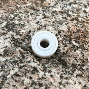 Op de afbeelding: Een witte keramische ring met drie concentrische cirkels. De ring ligt op een ruw, grijs, getextureerd oppervlak.