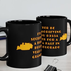 May include: Two black coffee mugs with a white interior. The mugs have a black handle and feature a yellow silhouette of a bulldozer with the text "HER BE I'M MODIFYING SECRET SU D355A ER FOR A KONA HALF IN BULCOLORADO YEAR GRAND" printed on the front.