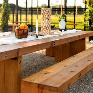 May include: A long, rectangular wooden table with a matching bench. A black wire basket holds oranges and lemons. A white runner, a candle, and a decorative lantern sit on the table. The table is outdoors.