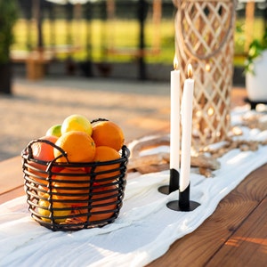 May include: An outdoor table arrangement with a black wire basket of oranges, lemons, and limes. Two lit white candles in black holders and a woven lantern are on a white runner. The table is made of wood.