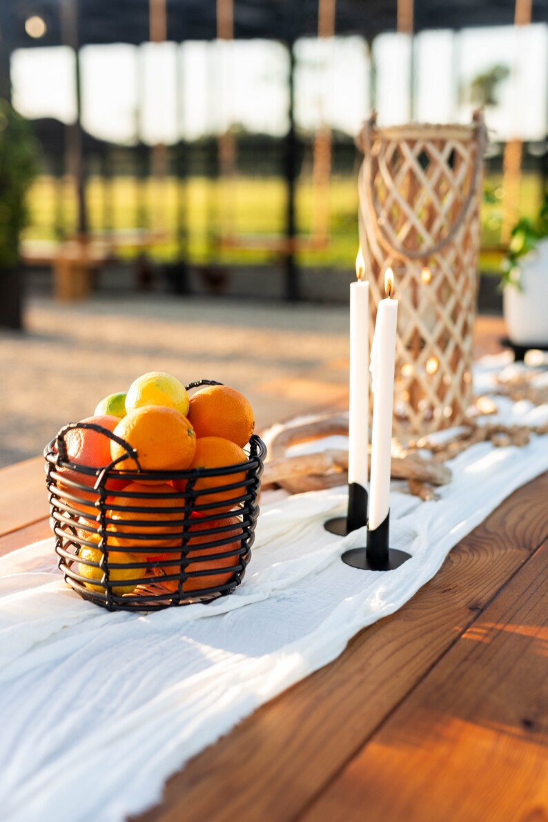 May include: An outdoor table arrangement with a black wire basket of oranges, lemons, and limes. Two lit white candles in black holders and a woven lantern are on a white runner. The table is made of wood.