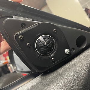 May include: A black car speaker with the Infinity logo, mounted on a black plastic panel. The speaker is secured with multiple screws and is part of a car's interior.
