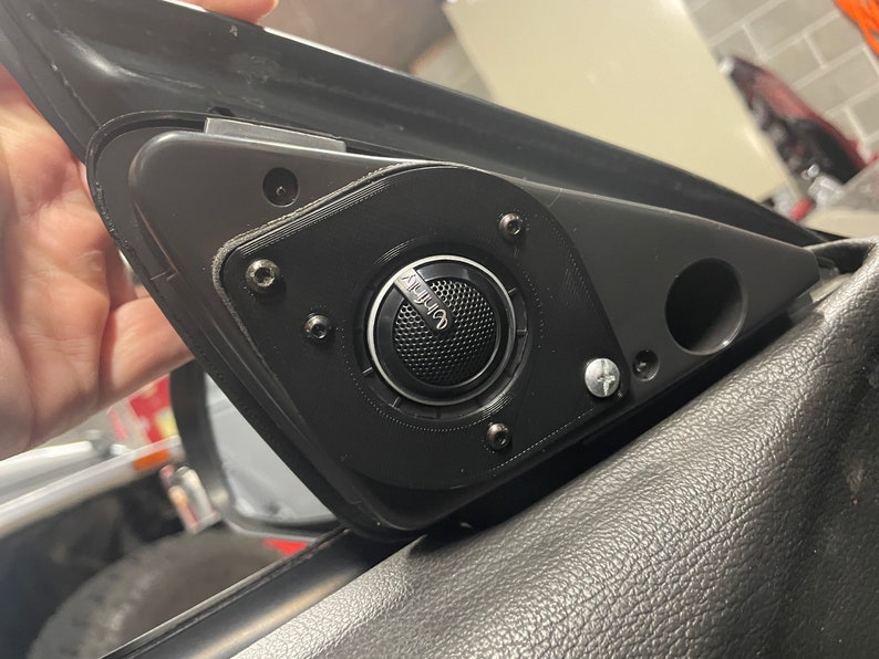 May include: A black car speaker with the Infinity logo, mounted on a black plastic panel. The speaker is secured with multiple screws and is part of a car's interior.