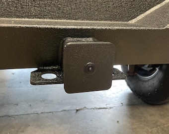 May include: A black square-shaped trailer hitch with a small black camera lens in the center.