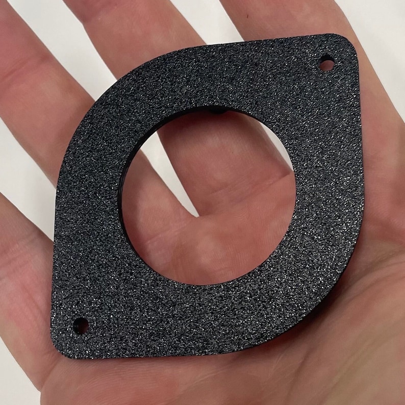 May include: A black, textured, geometric object with a circular opening and two small holes. The object has a unique shape, resembling a rounded square with a circle cut out of the center. The surface has a speckled appearance.