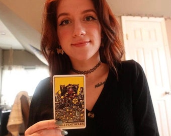 Könnte beinhalten: Eine rechteckige Tarotkarte mit der Abbildung des Königs der Münzen auf gelbem Hintergrund, einer auf einem Thron sitzenden Figur und dem unten in Schwarz aufgedruckten Text KING OF PENTACLES, gehalten vor einem schwarzen Langarmshirt.