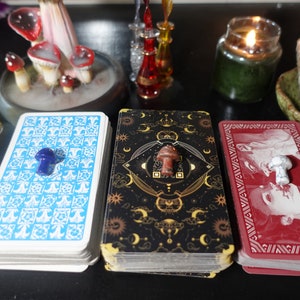 May include: Three decks of tarot cards with different coloured backs, each topped with a small mushroom figurine. The decks are blue, black, and red.