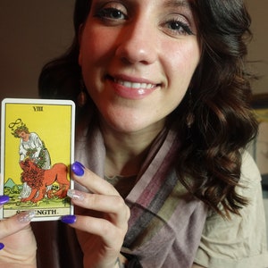 May include: A woman holds up a tarot card with the number VIII and the word STRENGTH printed on it. The card depicts a woman with a crown of flowers gently taming a lion.
