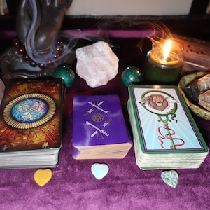 May include: Three tarot cards laid out on a purple velvet surface. The cards are surrounded by crystals, a burning candle, and smudging sticks. The cards are "The World", "Two of Swords", and "The Emperor".