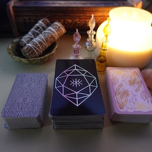 May include: Three decks of tarot cards on a table with a lit candle, sage bundles, and bottles of essential oils. The tarot decks have different designs, including a black deck with a geometric eye design, a purple deck with a textured pattern, and a pink deck with a floral design.