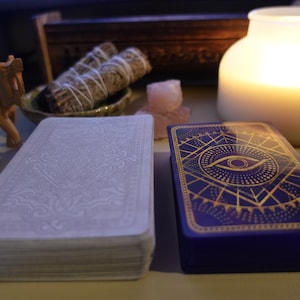 May include: Two decks of tarot cards, one with a white background and intricate designs, and the other with a blue background and a gold eye design. The cards are on a table with a candle and other decorative items.