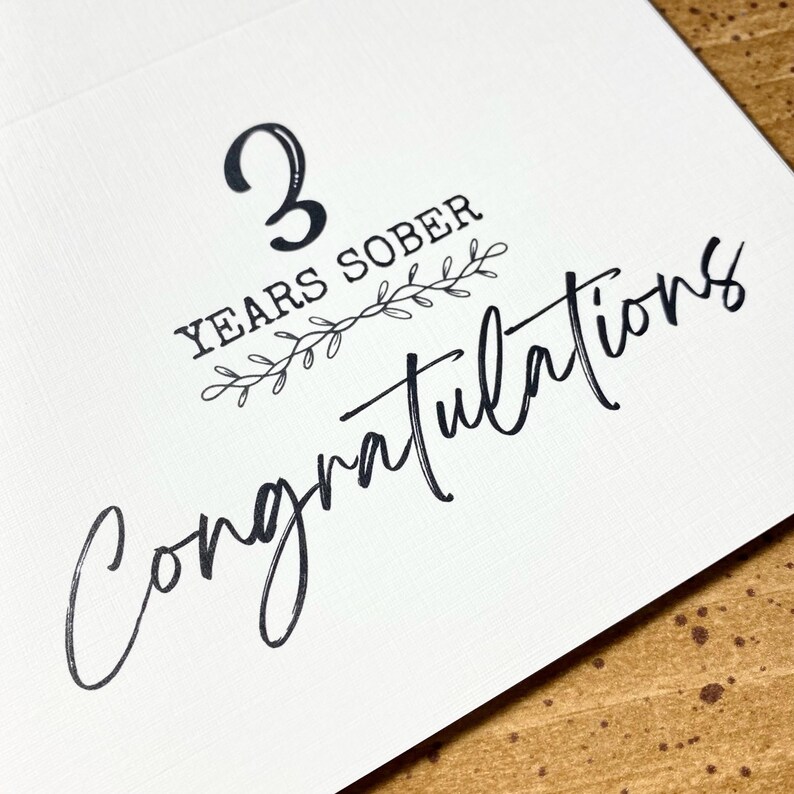 May include: A white card with black text that reads "3 Years Sober Congratulations". The text is in a handwritten font and is surrounded by a small floral design.
