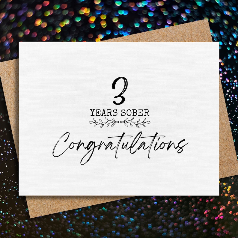 May include: A white card with black text that reads "3 Years Sober Congratulations". The card is decorated with a small floral design.