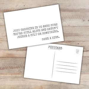 May include: A white rectangular postcard featuring black typewriter-style text that reads: JUST CHECKING IN TO MAKE SURE YOU'RE STILL ALIVE AND HAVEN'T JOINED A CULT OR SOMETHING. SEND A SIGN. The reverse side shows standard postcard lines and a stamp box.