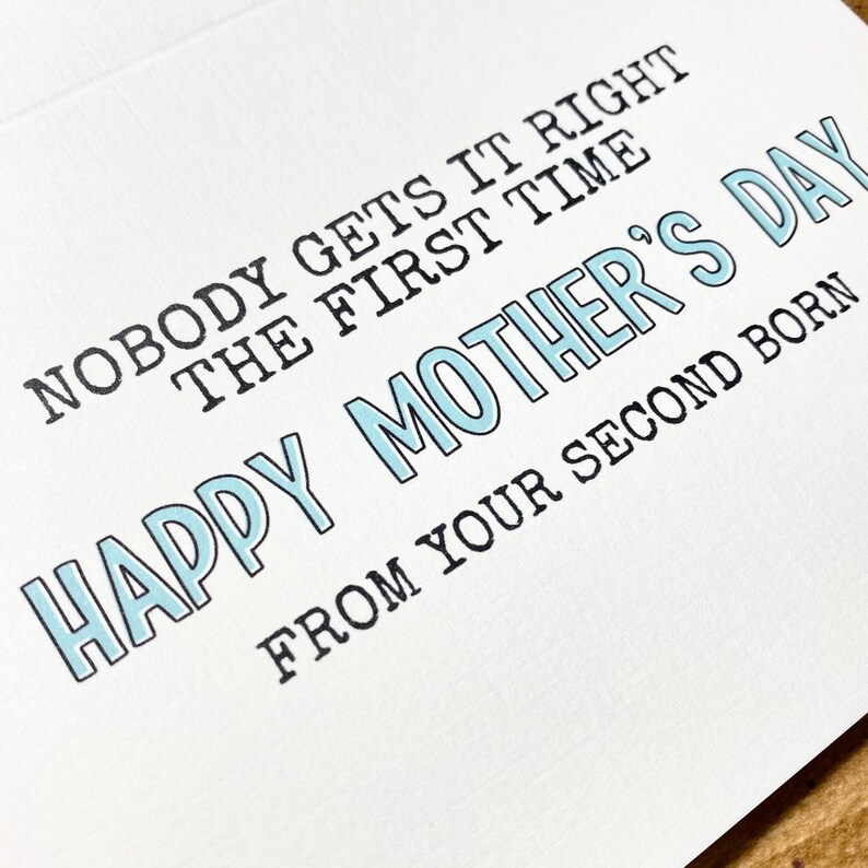 May include: A white greeting card with the text "NOBODY GETS IT RIGHT THE FIRST TIME HAPPY MOTHER'S DAY FROM YOUR SECOND BORN" in black and light blue lettering. The card is blank inside.