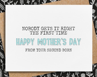 Puede incluir: Tarjeta blanca del Día de la Madre con el texto "NOBODY GETS IT RIGHT THE FIRST TIME HAPPY MOTHER'S DAY FROM YOUR SECOND BORN." La tarjeta está sobre un sobre marrón, sobre un fondo floral en blanco y negro.
