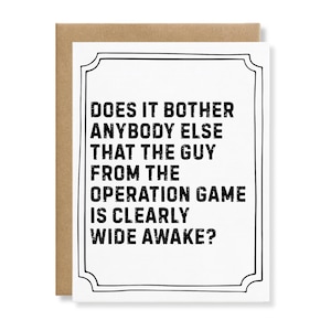 May include: A white greeting card with a black decorative border and the text: DOES IT BOTHER ANYBODY ELSE THAT THE GUY FROM THE OPERATION GAME IS CLEARLY WIDE AWAKE? The card is positioned against a plain brown envelope background.