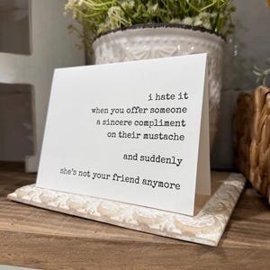 May include: A white greeting card with black typewriter-style text that reads: i hate it when you offer someone a sincere compliment on their moustache and suddenly she's not your friend anymore. The card is displayed on a white textured base.